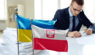 Mężczyzna podpisuje dokumenty, a obok niego znajdują się flagi Ukrainy i Polski