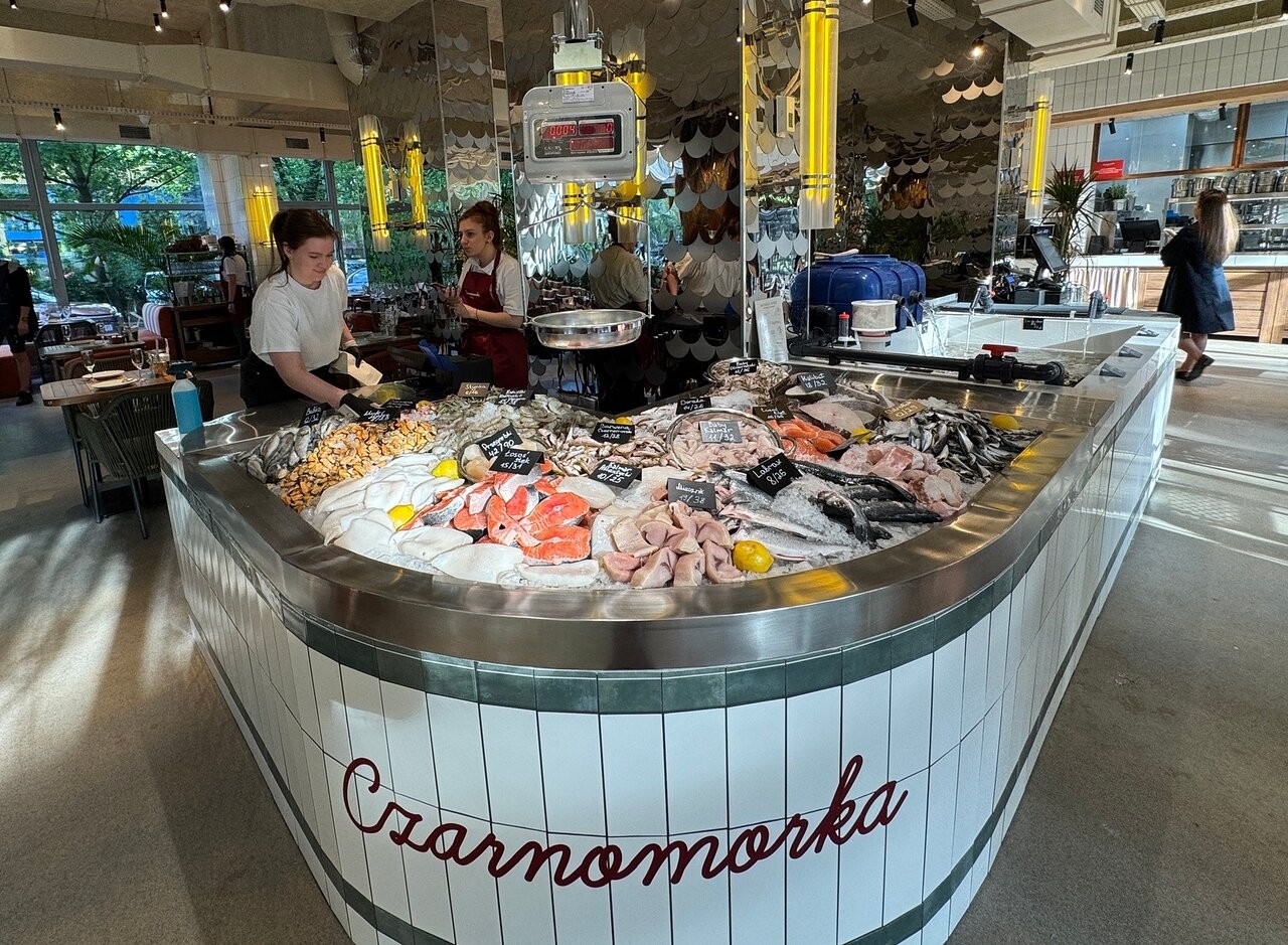 Жвавий рибний прилавок у ресторані, навколо якого зібралося кілька людей, які розглядають асортимент свіжих морепродуктів.